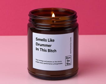 May include: A brown glass candle jar with a white label that reads "Smells Like Drummer In This Bitch". The label also includes the text "Your energy and passion on the drums make every performance unforgettable." and the logo "Burn The Wax" with the text "Hand Poured Soy Candle".