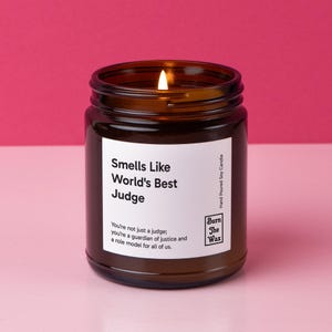 May include: A brown glass candle jar with a white label that reads "Smells Like World's Best Judge". The label also includes the text "You're not just a judge: you're a guardian of justice and a role model for all of us." and the logo "Burn The Wax".