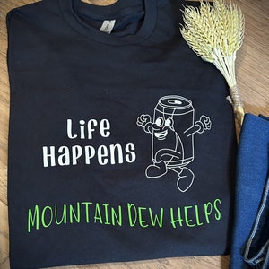 May include: Black t-shirt with white text that reads "Life Happens Mountain Dew Helps". A cartoon can of Mountain Dew is walking with a smile on its face.