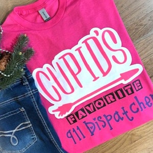 May include: Pink t-shirt with white text that says "Cupid's Favorite 911 Dispatcher". The text is in a blocky font and has an arrow pointing to the right.