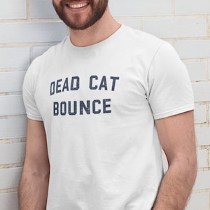 Może przedstawiać: Biała koszulka z nadrukiem "DEAD CAT BOUNCE" w kolorze niebieskim na przodzie. Osoba w koszulce stoi przed białą ceglaną ścianą.