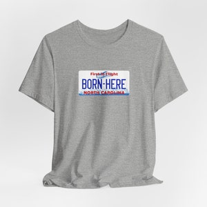 May include: Gray t-shirt with a blue and red North Carolina license plate graphic that says "First in Flight" and "Born-Here".