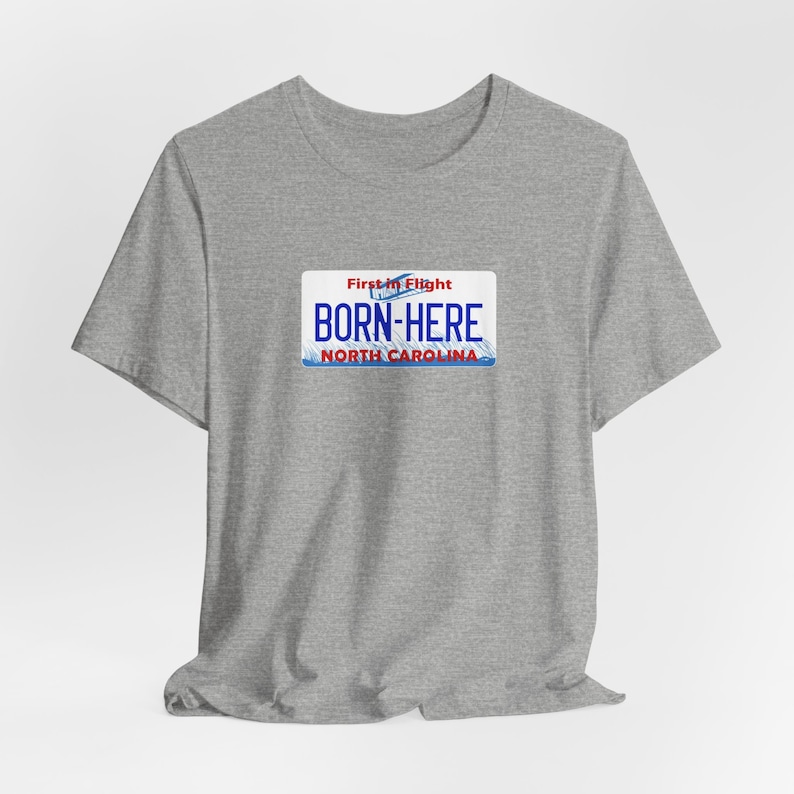 May include: Gray t-shirt with a blue and red North Carolina license plate graphic that says "First in Flight" and "Born-Here".