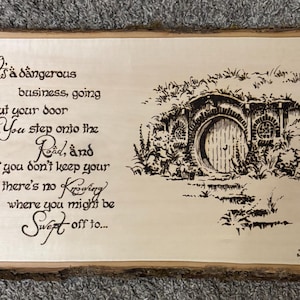 May include: Wooden sign with a natural edge, featuring a pyrography design. The artwork includes a hobbit hole illustration and the quote, "It's a dangerous business, going out your door..." The sign is a light tan color.
