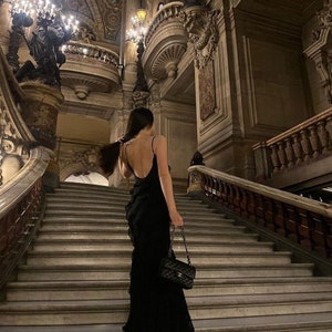 Puede incluir: Una mujer con un vestido negro con volantes se encuentra en una gran escalera en un edificio hist&oacute;rico. Lleva un bolso negro con una correa de cadena.