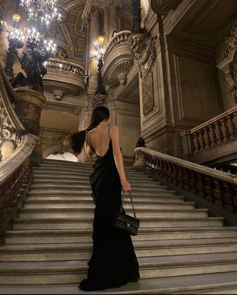 Puede incluir: Una mujer con un vestido negro con volantes se encuentra en una gran escalera en un edificio hist&oacute;rico. Lleva un bolso negro con una correa de cadena.