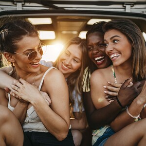 May include: Four women laughing together in the back of a car. They are all smiling and looking at the camera. The woman in the front has her arms around the woman behind her.