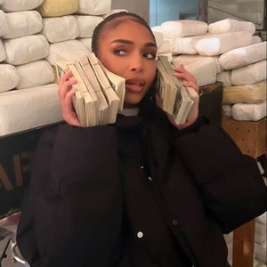 Puede incluir: Una persona con una chaqueta negra y pantalones vaqueros sostiene fajos de dinero en efectivo a cada lado de la cabeza. El fondo muestra una pared con el texto "The Plug" y una pila de objetos rectangulares blancos.