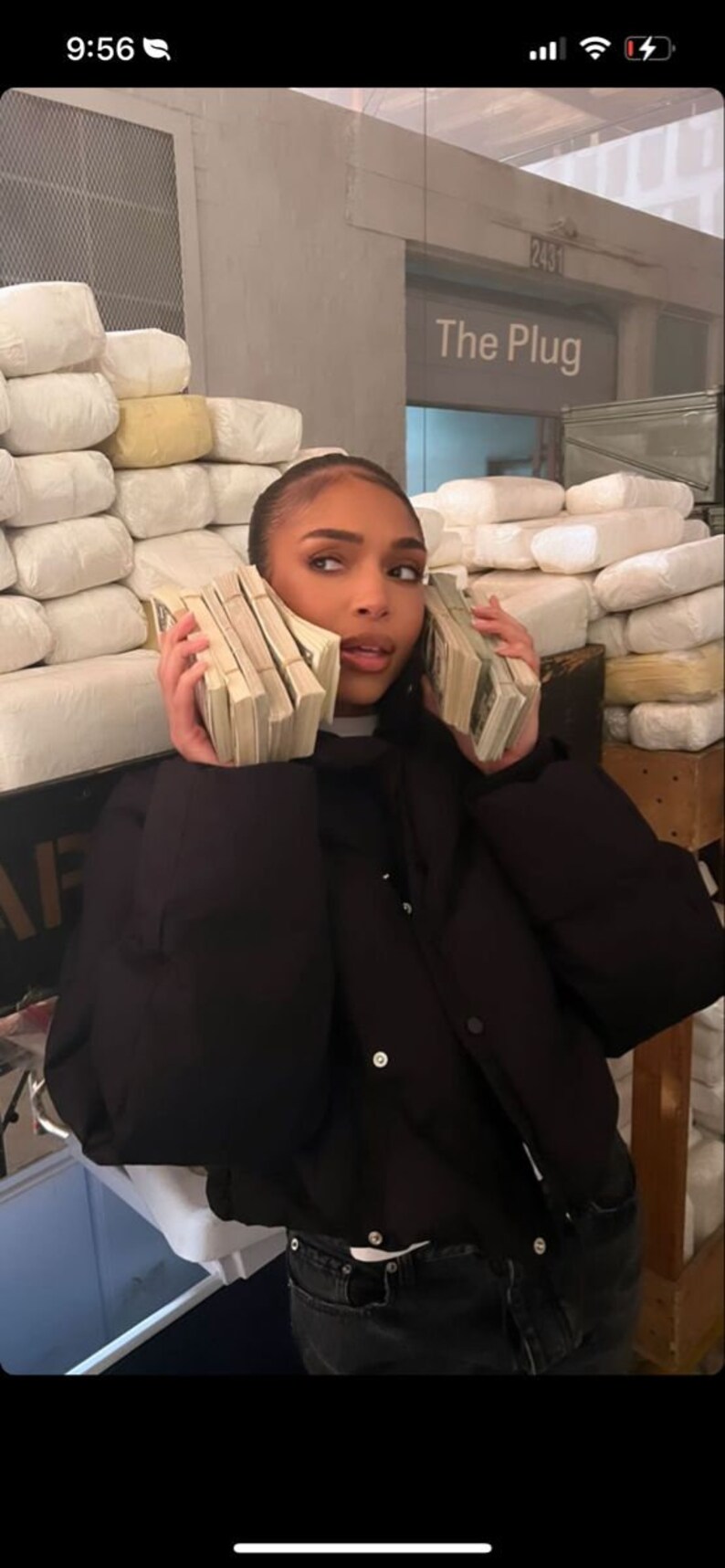 Puede incluir: Una persona con una chaqueta negra y pantalones vaqueros sostiene fajos de dinero en efectivo a cada lado de la cabeza. El fondo muestra una pared con el texto "The Plug" y una pila de objetos rectangulares blancos.
