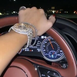 Puede incluir: Primer plano del volante de un coche con un interior de cuero marr&oacute;n. El volante tiene un logotipo de Bentley plateado y una pantalla digital que muestra la velocidad del coche y otra informaci&oacute;n. Una mano con un reloj y una pulsera engastados con diamantes agarra el volante.