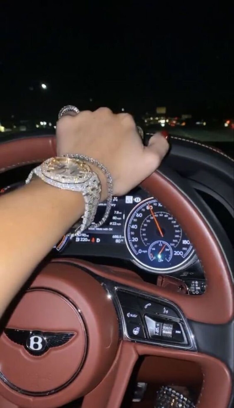 Puede incluir: Primer plano del volante de un coche con un interior de cuero marr&oacute;n. El volante tiene un logotipo de Bentley plateado y una pantalla digital que muestra la velocidad del coche y otra informaci&oacute;n. Una mano con un reloj y una pulsera engastados con diamantes agarra el volante.