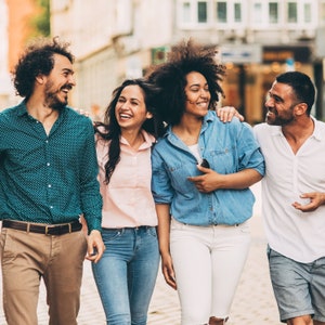 May include: Four friends, two women and two men, are walking down a city street and laughing together. They are all wearing casual clothing.