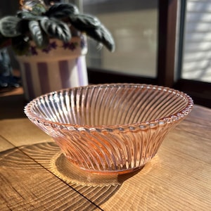 May include: A vintage pink glass bowl with a spiral design. The bowl has a scalloped rim and sits on a wooden surface. The bowl is likely used for serving or display.