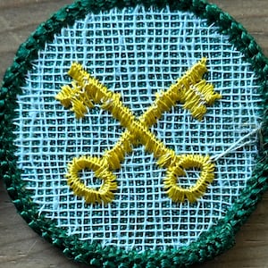 Op de afbeelding: Geborduurde patch met een lichtblauwe rasterachtergrond en een groene rand. Twee gele sleutels zijn in het midden gekruist. De patch is rond van vorm.