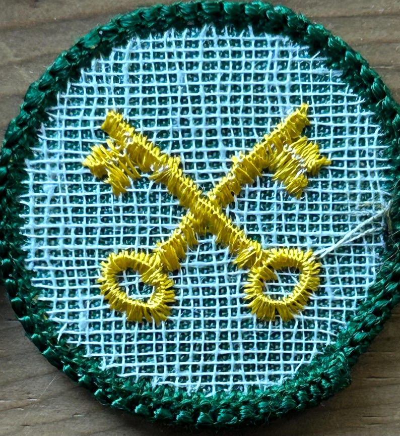 Op de afbeelding: Geborduurde patch met een lichtblauwe rasterachtergrond en een groene rand. Twee gele sleutels zijn in het midden gekruist. De patch is rond van vorm.