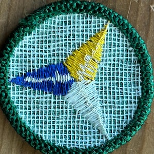 Op de afbeelding: Geborduurde patch met een groene rand en een witte mesh-achtergrond. De patch heeft een blauw-wit ontwerp met een gele accent, dat lijkt op een gestileerde pijl of embleem. De patch is rond.
