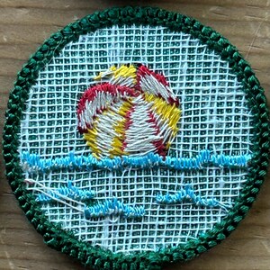 Op de afbeelding: Geborduurde patch met een zonsondergang boven water. De zon is afgebeeld in geel, rood en wit, met blauwe golven eronder. De patch heeft een groene rand en een witte mesh-achtergrond.