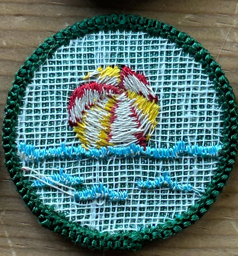 Op de afbeelding: Geborduurde patch met een zonsondergang boven water. De zon is afgebeeld in geel, rood en wit, met blauwe golven eronder. De patch heeft een groene rand en een witte mesh-achtergrond.