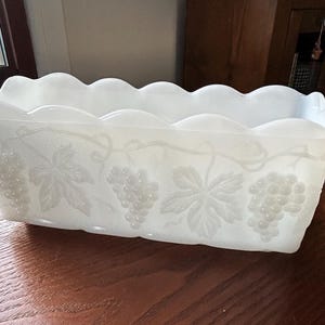 May include: A rectangular, white, frosted glass dish with a scalloped rim. The sides are decorated with embossed grape clusters and leaves. The dish is sitting on a wooden table.