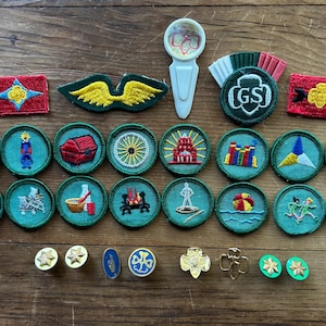 Op de afbeelding: Een verzameling Girl Scout-badges en -spelden, waaronder ronde, geborduurde patches met verschillende ontwerpen, een groen en wit GS-embleem en goudkleurige spelden. De badges tonen afbeeldingen van een tent, een schatkist en meer. De items zijn gerangschikt op een houten oppervlak.