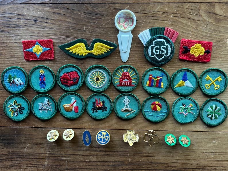Op de afbeelding: Een verzameling Girl Scout-badges en -spelden, waaronder ronde, geborduurde patches met verschillende ontwerpen, een groen en wit GS-embleem en goudkleurige spelden. De badges tonen afbeeldingen van een tent, een schatkist en meer. De items zijn gerangschikt op een houten oppervlak.