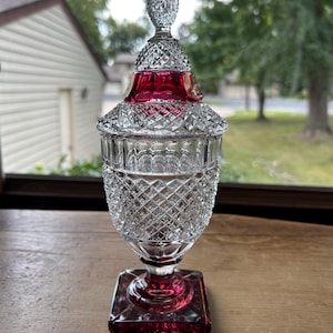 May include: A clear and ruby red glass candy dish with a lid. The piece features a square base, a pedestal stem, and a body decorated with a diamond cut pattern. The lid is topped with a pointed finial and includes a band of ruby red glass.
