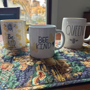 May include: Three ceramic mugs with bee-themed designs. One mug is yellow with a honeycomb pattern and the words "Be Kind". Another mug is white with "Bee Kind" and a bee graphic. The third mug is white with "Queen" and a bee.