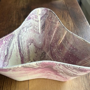 May include: A decorative, triangular-shaped bowl with a marbled design. The bowl features shades of pink, white, and gray, creating a swirling pattern. The bowl is placed on a wooden surface.