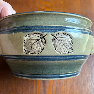 May include: A ceramic bowl with a green and blue glaze. Two leaf imprints are centered on the bowl's exterior. The bowl has a wide rim and two small handles. The bowl is approximately 6 inches in diameter.