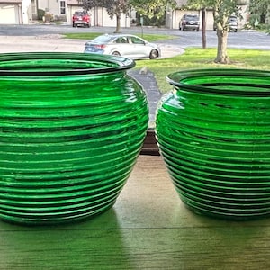 May include: Two vibrant green glass bowls featuring a distinct horizontal ribbed texture. Both vessels have a wide, rounded body shape with a smooth, flared rim at the top. The glass is translucent, allowing light to pass through the green material.