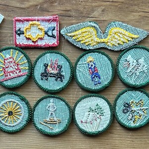 Op de afbeelding: Een verzameling vintage geborduurde patches in verschillende vormen en ontwerpen. De patches tonen afbeeldingen van een kompas, een kampvuur, een schip en andere symbolen. Het kleurenpalet omvat groen, rood, geel en wit.