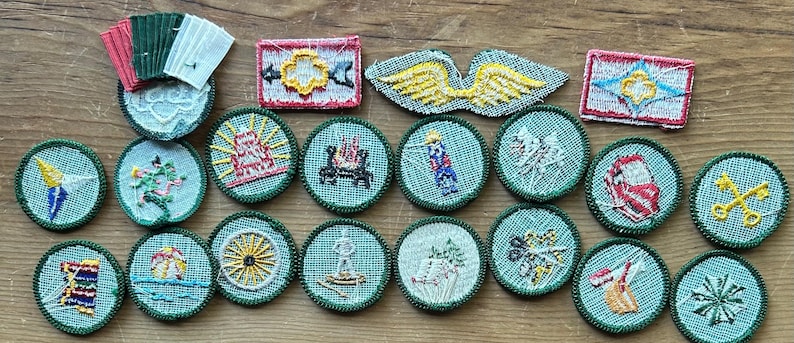 Op de afbeelding: Een verzameling vintage geborduurde patches in verschillende vormen en ontwerpen. De patches tonen afbeeldingen van een kompas, een kampvuur, een schip en andere symbolen. Het kleurenpalet omvat groen, rood, geel en wit.