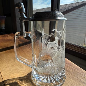 May include: A clear glass beer stein featuring an etched design of a hunting dog in a field with birds in flight. The stein has a metal lid with a decorative border and a metal handle topped with a small sculpted dog figurine.