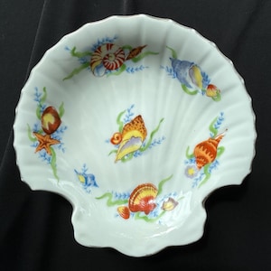 May include: A white porcelain dish shaped like a scallop shell with a scalloped edge and a silver-toned rim. The interior features hand-painted illustrations of various seashells, including conch and starfish, interspersed with green seaweed.