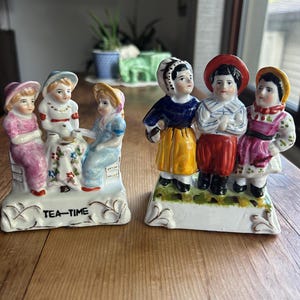 May include: Two porcelain figurines on rectangular bases. The left piece features three figures seated at a table with the text TEA-TIME. The right piece features three standing figures wearing colorful traditional-style clothing and hats.