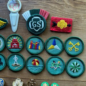 Op de afbeelding: Een verzameling Girl Scout-badges en -spelden, waaronder ronde geborduurde patches met verschillende ontwerpen en een speld met de letters "GS". De badges tonen afbeeldingen van een vuurtoren, een strandbal en andere symbolen.