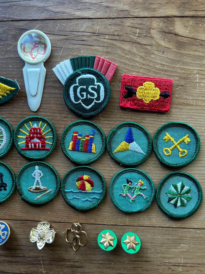 Op de afbeelding: Een verzameling Girl Scout-badges en -spelden, waaronder ronde geborduurde patches met verschillende ontwerpen en een speld met de letters "GS". De badges tonen afbeeldingen van een vuurtoren, een strandbal en andere symbolen.