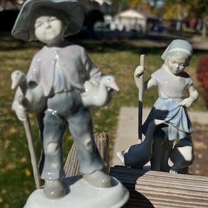 May include: Two porcelain figurines featuring a person in a wide-brimmed hat holding a goose and a staff, and a second person in a headscarf holding a staff with two geese. Both figures are painted in soft shades of blue, gray, and white.