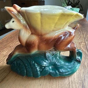 May include: A ceramic planter shaped like a deer with a large, fan-like shell structure on its back. The deer is colored in shades of brown and cream, while the base is a deep, glossy green with molded leaf details and a smooth, glazed finish.
