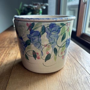 May include: A cylindrical ceramic planter with a cream-colored base featuring a hand-painted pattern of blue flowers, green leaves, and small pink accents. The rim has a speckled blue finish, and the vessel has a smooth, slightly tapered shape.