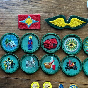 Op de afbeelding: Een verzameling vintage geborduurde patches en spelden. De patches hebben verschillende ontwerpen, waaronder een rode rechthoek met een ster, groene vleugels en ronde patches met kampeer-, kaars- en wagenwielmotieven. De spelden zijn stervormig en ovaal.