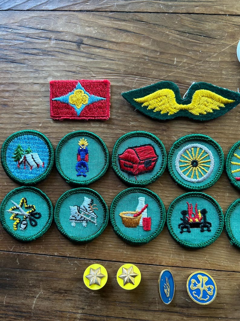 Op de afbeelding: Een verzameling vintage geborduurde patches en spelden. De patches hebben verschillende ontwerpen, waaronder een rode rechthoek met een ster, groene vleugels en ronde patches met kampeer-, kaars- en wagenwielmotieven. De spelden zijn stervormig en ovaal.