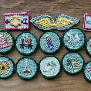 Op de afbeelding: Een verzameling vintage geborduurde patches in verschillende vormen en ontwerpen. De patches tonen afbeeldingen van vleugels, een kampvuur, een wagenwiel en andere symbolen, met een kleurenpalet van groen, rood, geel en wit. De patches zijn gerangschikt op een houten oppervlak.