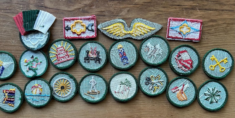 Op de afbeelding: Een verzameling vintage geborduurde patches in verschillende vormen en ontwerpen. De patches tonen afbeeldingen van vleugels, een kampvuur, een wagenwiel en andere symbolen, met een kleurenpalet van groen, rood, geel en wit. De patches zijn gerangschikt op een houten oppervlak.