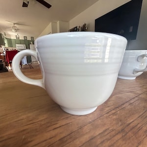 May include: A white ceramic mug featuring a rounded body with horizontal ribbed detailing around the upper portion, a curved handle on the side, and a small pedestal base. The mug has a smooth, glossy finish and a wide, open top rim.