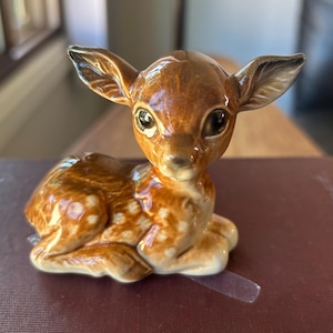 May include: A glossy ceramic figurine of a young deer in a resting position. The fawn features a rich brown coat with white spots along its back and sides, large dark eyes, and upright ears, finished with a smooth, reflective glazed surface.