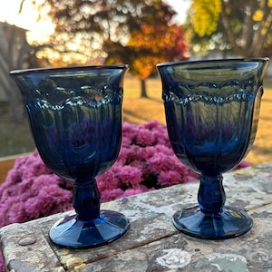 May include: Two cobalt blue glass goblets with a fluted design. The goblets are sitting on a wooden surface with a blurred background of pink flowers.