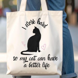 以下が含まれることがあります： 黒い猫のシルエットと「I work hard so my cat can have a better life」の文字が入ったクリーム色のトートバッグ。デザインには2つの小さなピンクのハートが含まれています。バッグには白いハンドルが付いています。