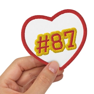 May include: A red heart-shaped patch with a white background and yellow embroidered text that says "#87".