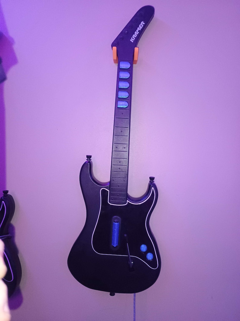 Op de afbeelding: Zwarte Kramer gitaarvormige gamecontroller die aan een muur hangt. De controller heeft blauwe en witte accenten, met de merknaam "Kramer" op de kop. De controller is gemonteerd met oranje beugels.